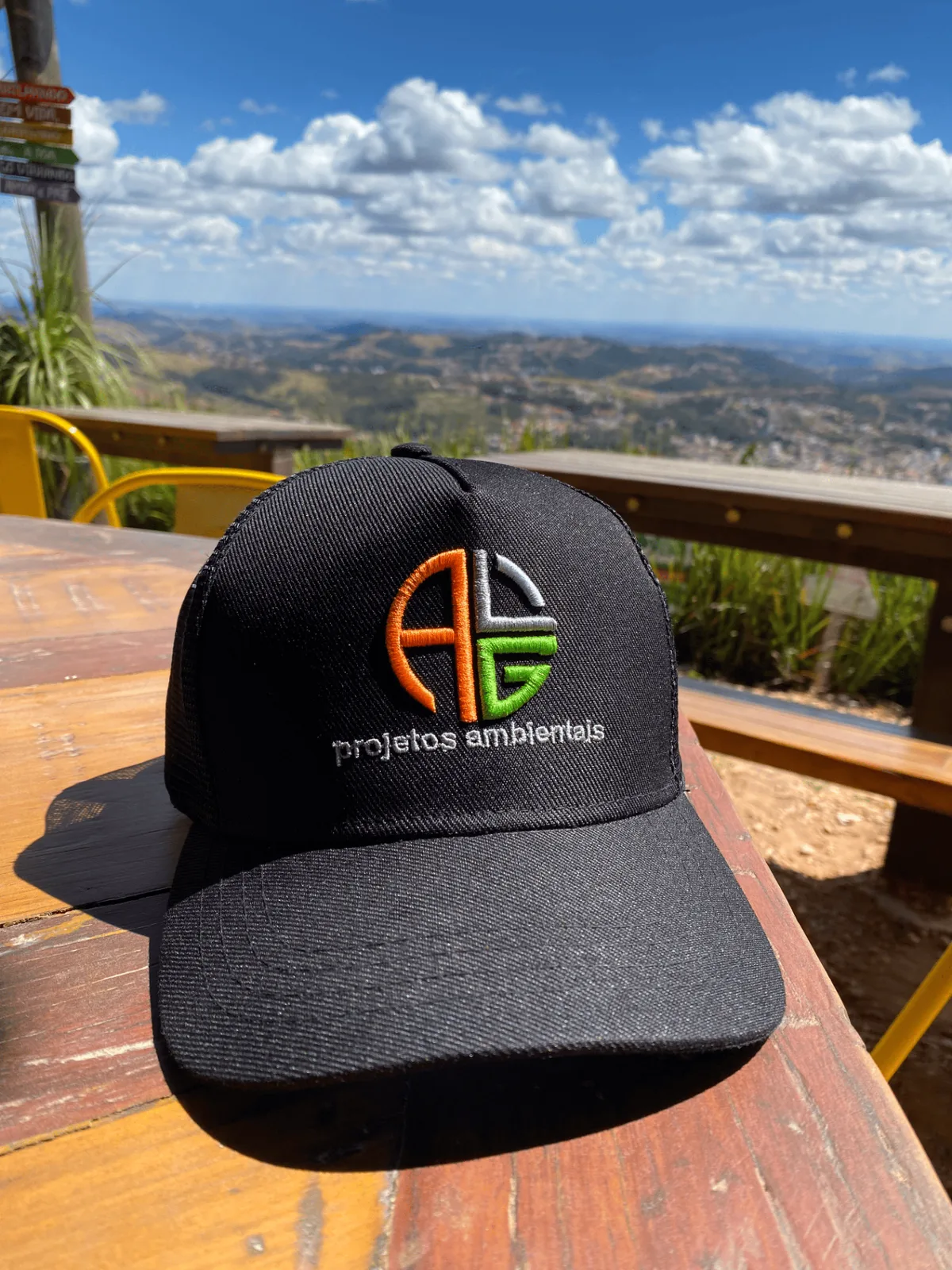 Boné da ALG Projetos Ambientais sobre mesa de madeira com paisagem de serra ao fundo